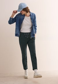 Denim bomberjacka, vit t-shirt, mörkgröna byxor med dragsnöre, vita sneakers och ljusblå keps. Avslappnad outfit med minimal detalj.