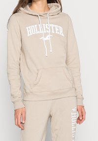 Žena v béžové mikině Hollister s bílými stahovacími šňůrkami a odpovídajících teplákách, které mají bílou logovku Hollister na stehně.