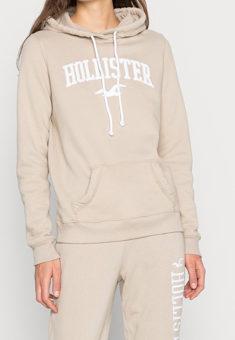 Žena v béžové mikině Hollister s bílými stahovacími šňůrkami a odpovídajících teplákách, které mají bílou logovku Hollister na stehně.