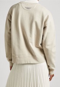 Sudadera color beige con un corte relajado, cuello de canalé y un pequeño diseño decorativo encima del cuello en la parte posterior. Tejido suave y texturizado.