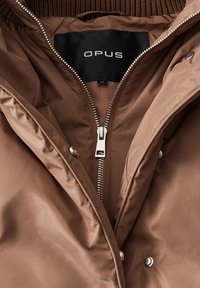 Giacca marrone imbottita con colletto a coste; presenta una zip argentata e una chiusura a scatto. L'etichetta mostra "OPUS" in lettere bianche e in grassetto.
