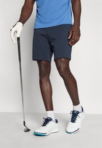 Γκολφ σορτς σε σκούρο μπλε ύφασμα, φανελάκι με εφαρμοστή γραμμή σε μπλε χρώμα, λευκά και μπλε παπούτσια γκολφ και μια ράβδος γκολφ που κρατιέται σε χέρι με γάντι.