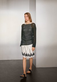 Schwarzer Strickpullover mit langen Ärmeln, über einem weißen Satinkleid mit schwarzem Spitzenrand getragen. Schwarze flache Sandalen runden das Outfit ab.