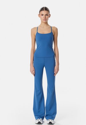 Žena stojaca čelom vpred, oblečená v modrý priliehavý tielko, zladené modré zvonové nohavice a biele tenisky na jednoliatom pozadí.