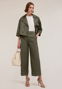 Olijfgroen kort jasje met dubbele knopen, gecombineerd met hoge olijfgroene wijde broek. Witte blouse eronder, met een crèmekleurige tas.