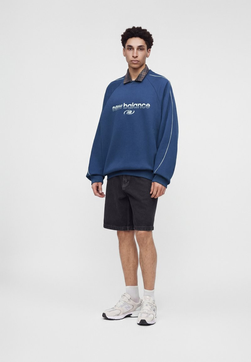 Νέος άντρας με μπλε φούτερ New Balance, μαύρες βερμούδες, λευκά αθλητικά παπούτσια και λευκές κάλτσες, στέκεται μπροστά σε ένα μονόχρωμο γκρι φόντο.