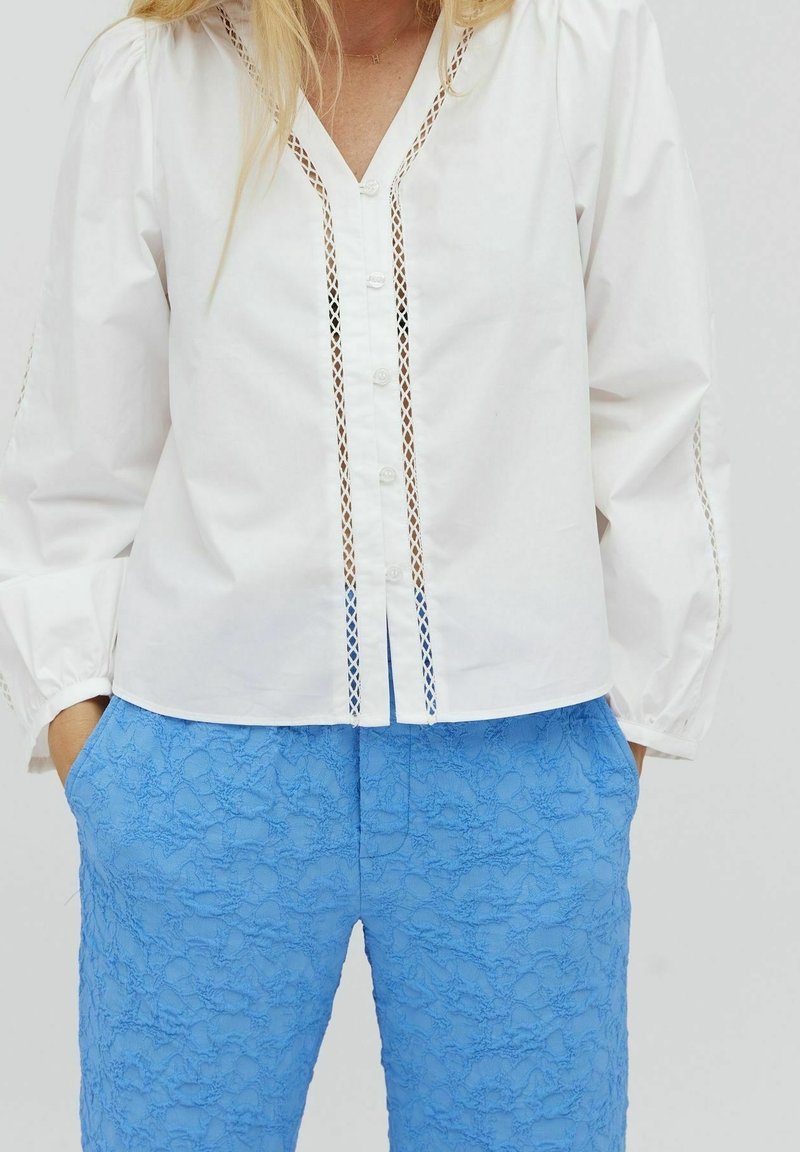 Camicia bianca con scollo a V e dettagli decorativi a taglio; abbinata a pantaloni leggeri blu chiaro con texture. La blusa ha maniche arricciate.