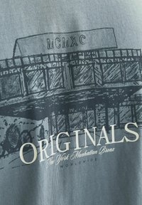 Tricou gri din bumbac cu un desen în negru al unei clădiri și textul "MCMXC" și "ORIGINALS" cu font alb îngroșat. Textul include detalii despre locație.