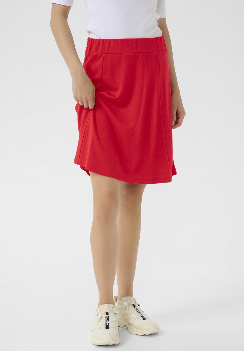 Jupe rouge avec une taille élastique, au design longueur genoux et texture lisse. Portée avec des chaussures de sport de couleur claire.