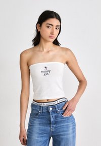 Fehér, pánt nélküli felső, amelyen a "tommy girl" szöveg és csillag logó látható. Világoskék farmernadrággal párosítva, amelyen márkás derékpánt található. Simaszerkezetű anyag.