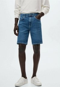 Shorts en denim de teinte bleu moyen avec une coupe droite, cinq poches et une fermeture à bouton. Associés à des baskets blanches et une chemise à manches longues légère.
