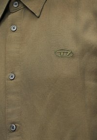 Πράσινο ελιάς πουκάμισο με γιακά, τέσσερα κουμπιά και ένα μικρό κεντητό οβάλ λογότυπο στην αριστερή πλευρά του στήθους.