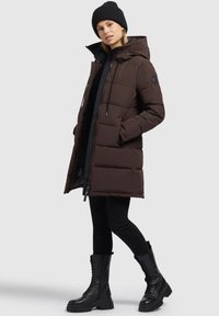 Bruine gewatteerde parka met capuchon, trekkoord en zijzakken, gedragen over zwarte leggings en zwarte veterschoenen.
