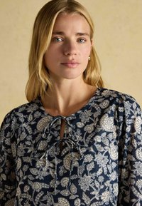 Navyblaue Bluse mit weißen Blumenmustern, runder Ausschnitt, gebundenes Detail an der Vorderseite und Puffärmeln aus leichtem Stoff.