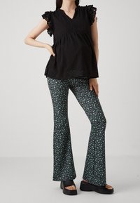 Blouse noire à volants avec un décolleté en V, associée à un pantalon évasé à motifs floraux en vert et noir. Présente un motif texturé sur la blouse.