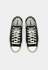 Ténis de lona pretos com atacadores brancos e solas de borracha. Apresentam biqueira e remendos com o logótipo "CONVERSE ALL STAR" na língua.