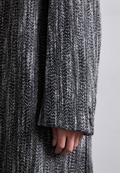 Manteau texturé noir et blanc avec un motif à côtes verticales, comportant un poignet à boutons et un gros plan de la manche.