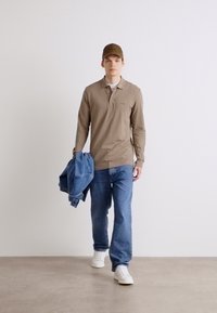 Polo de manga longa castanho claro, jeans de ganga azuis, sapatilhas brancas e um boné castanho a condizer, com uma jaqueta de ganga pendurada a um braço.