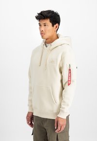 Krēmpauru krāsas kapucīna ar mīkstu audumu, priekšējo kabatu, kapuci ar auklu un sānu kabatu, kurā ir sarkans uzlīme. Redzama detalizēta šūšana.