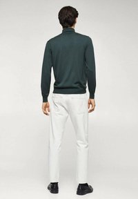 Tumma vihreä neule, jossa on korkea kaulus, kapea malli ja pitkät hihat, yhdistettyinä valkoisiin farkkuihin ja mustiin kenkiin. Tekstuuri näyttää sileältä.