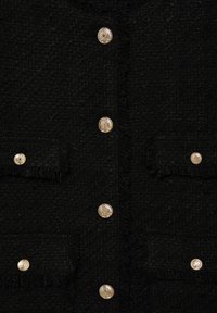 Mustekallinen musta kangas, jossa on kuusi kultavaahtopainiketta järjestetty pystysuoraan ja kaksi taskua, jotka on jokainen varmistettu kultanapilla.