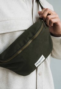 Man wearing light beige jacket holds dark green Holzhichter Berlin branded crossbody bag with gold zipper.