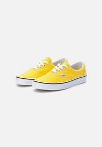 Gule kanvas sneakers med hvite snorer, gummisåler og en kontrasterende svart linje langs sålen, med et lite logomerke på siden.