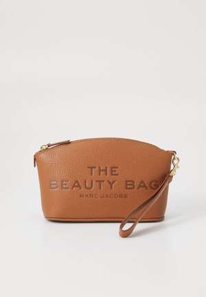 Brązowa skórzana kosmetyczka o teksturowanej powierzchni z złotym zamkiem, w kształcie zaokrąglonym, z wytłoczonym napisem "THE BEAUTY BAG" i "MARC JACOBS", z paskiem na nadgarstek.