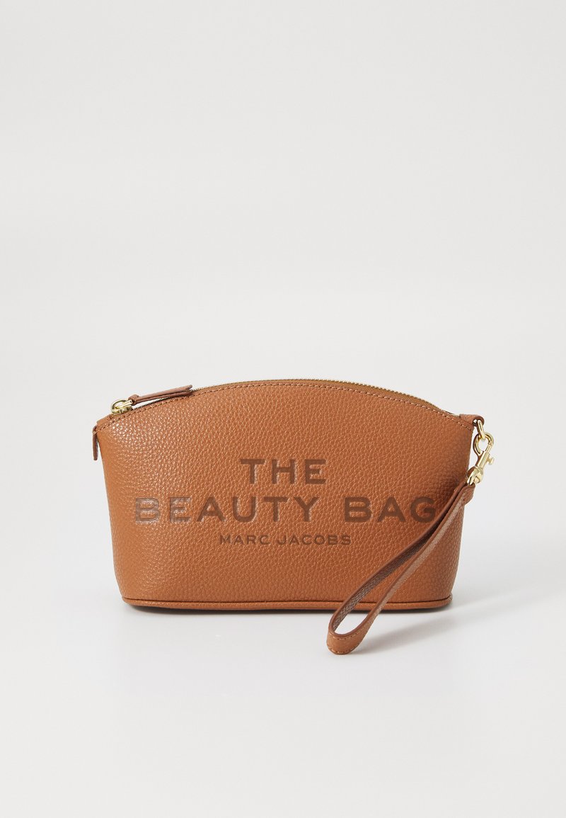 Bolsa de belleza de cuero marrón texturizado con cremallera dorada, forma redondeada, con letras en relieve "THE BEAUTY BAG" y "MARC JACOBS", que incluye una correa para la muñeca.