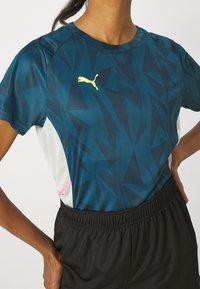 Kvinna som bär en blå mönstrad Puma sportskjorta med vita sidopaneler och svarta elastiska shorts, händerna på höfterna.