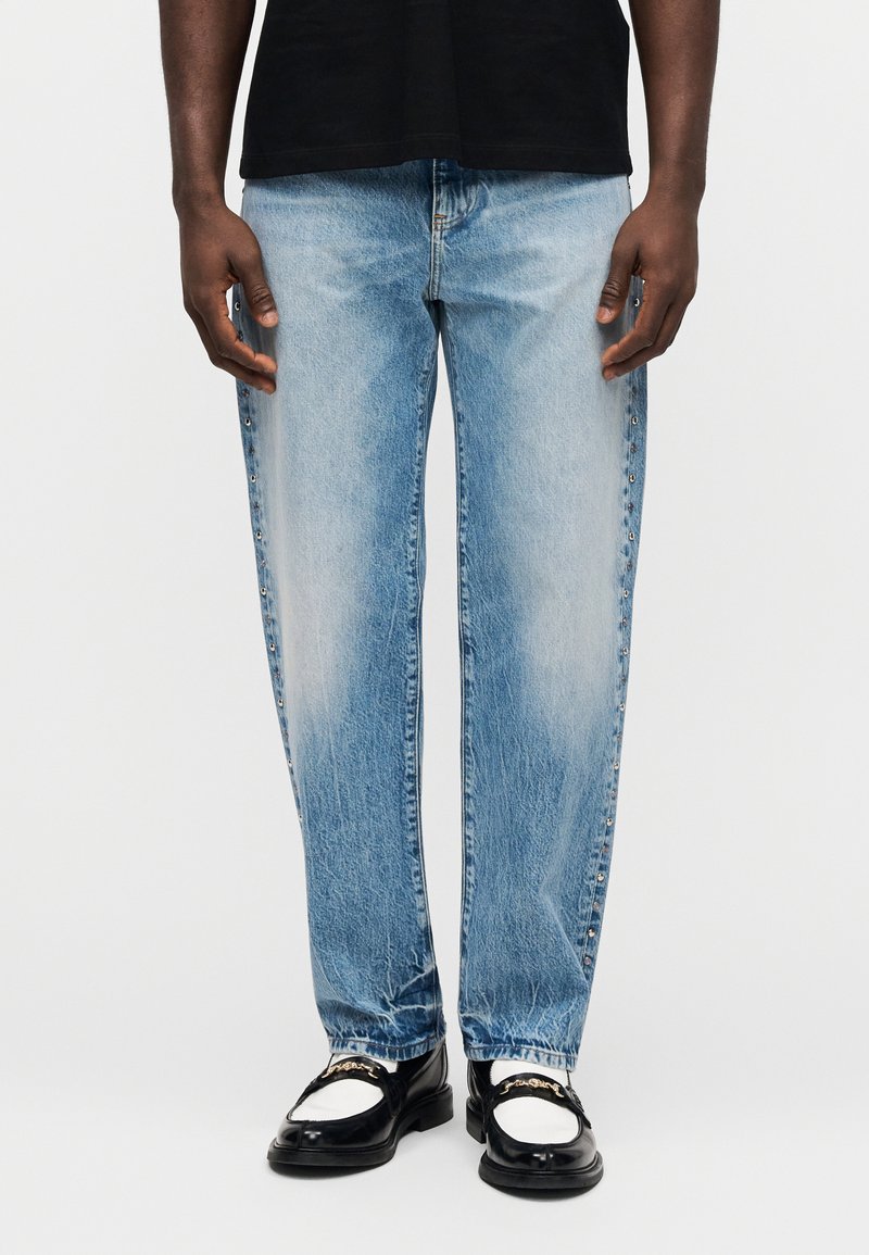 Personne portant un jean droit bleu clair avec des détails cloutés sur le côté, des mocassins noir et blanc, et un haut noir, debout devant un fond uni.