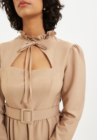 Beiges Langarmkleid mit Rüschenausschnitt, Herz-Ausschnitt und gebundenem Schleifchen. Es hat ein tailliertes Oberteil und einen Stoffgürtel mit quadratischer Schnalle.