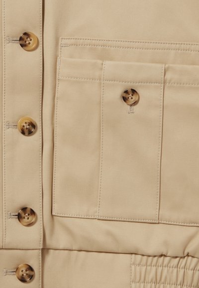 Vêtement en tissu beige avec de grands boutons marron, une poche poitrine et des détails élastiques sur la partie inférieure. Texture lisse et design ajusté.