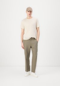 Beige linnen t-skjorte kombinert med olivengrønne bukser. Modellen har på seg en beige caps og hvite joggesko, stående mot en enkel hvit bakgrunn.
