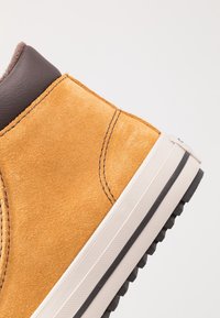 Zapatilla de ante amarilla con cuello de cuero marrón, suela de goma blanca y detalles de costura visibles en la parte superior. Acabado texturizado.