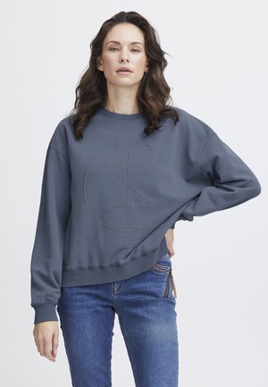 Sweatshirt gris foncé avec un col rond, des épaules tombantes et un design de cercle clouté à l'avant. Assorti avec un jean bleu.