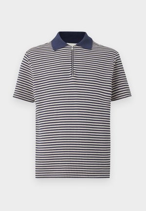 Mężczyzna w krótkim rękawie koszulki polo w granatowo-białe paski, czarnych spodniach i czarnych tenisówkach, stojący na tle gładkiego szarego tła.