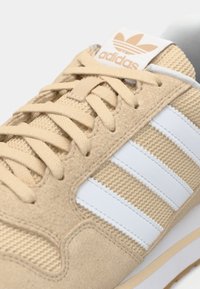 Chaussure de sport beige en suède et mesh. Comprend trois bandes blanches, des lacets beige et une languette blanche avec un logo en relief.