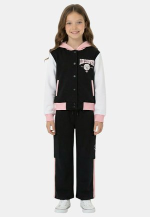 Jeune fille portant une veste universitaire noire et blanche avec des accents roses, un pantalon noir assorti avec des bandes roses et des baskets blanches.