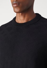 Homme portant un pull en maille texturée noire avec un col rond, visible du menton jusqu'au haut de la poitrine, sur un fond clair uni.