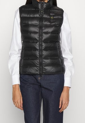 Frau trägt eine schwarze gesteppte ärmellose Weste über einem weißen Langarmshirt und dunkelblauen Jeans, steht vor einem einfarbigen Hintergrund.