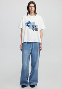 Witte katoenen t-shirt met een blauw bloemenpatroon en het woord "UNIQUE" in zwart. Gecombineerd met lichtblauwe wijde jeans.