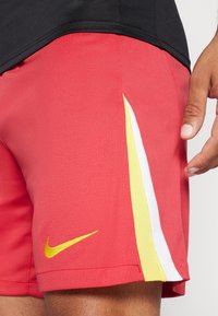 Punased sportlikud lühikesed püksid, millel on tekstuuriga kangas, kollased ja valged detailid külgedel ning väike kollane Nike logo hemmi lähedal.