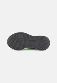 Semelle de chaussure en caoutchouc noir avec un profil texturé, présentant des motifs circulaires et rectangulaires, un talon courbé et des accents verts sur le côté.