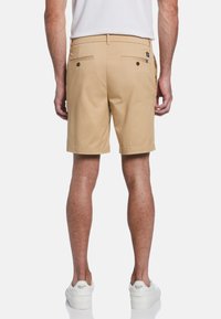 Shorts en coton beige avec une coupe droite, dotés de poches arrière et d'une petite étiquette logo. Portés avec des baskets blanches, mettant en valeur un design décontracté.