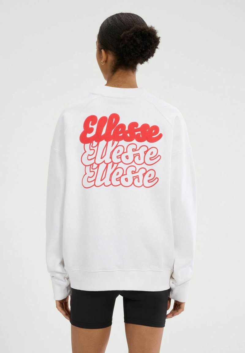 Mujer con cabello rizado que lleva una sudadera blanca con el logo rojo de "Ellesse" impreso tres veces en la parte de atrás, combinada con pantalones cortos negros.