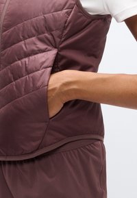 Αμάνικο μπουφάν σε βαθύ μοβ, με φερμουάρ εμπρός, πλαϊνή τσέπη και καπιτονέ σχέδιο. Λείο, μονωτικό ύφασμα.