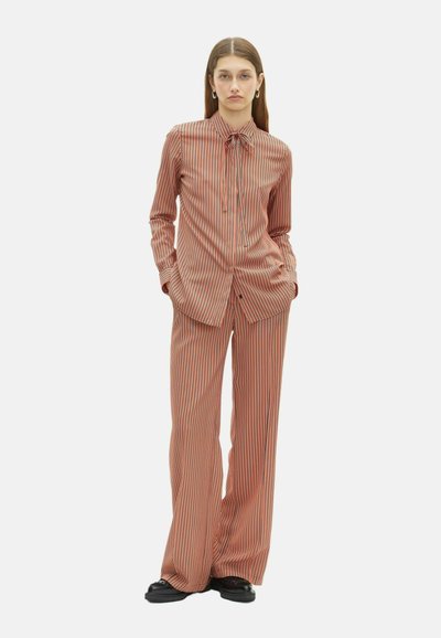 Vrouw in een loszittende oranje en zwarte verticaal gestreepte blouse met strik-kraag en bijpassende bredere broek, staand met haar handen in de zakken.