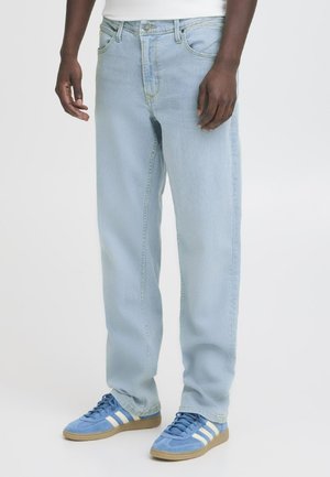 Person iført lyseblå straight-leg jeans og blå joggesko med hvite striper på en enkel hvit bakgrunn.