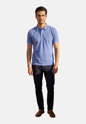 Uomo in piedi, indossa una polo azzurro chiaro a maniche corte, pantaloni scuri e mocassini marroni su uno sfondo chiaro e uniforme.